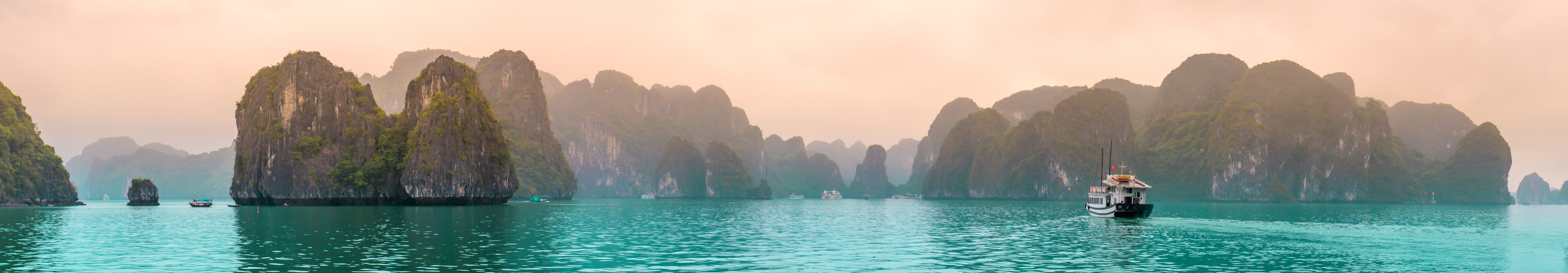Panorama der Halong-Bucht in Vietnam mit türkisfarbenem Wasser, Kalksteinfelsen und einem Boot im Vordergrund bei nebliger Abendstimmung