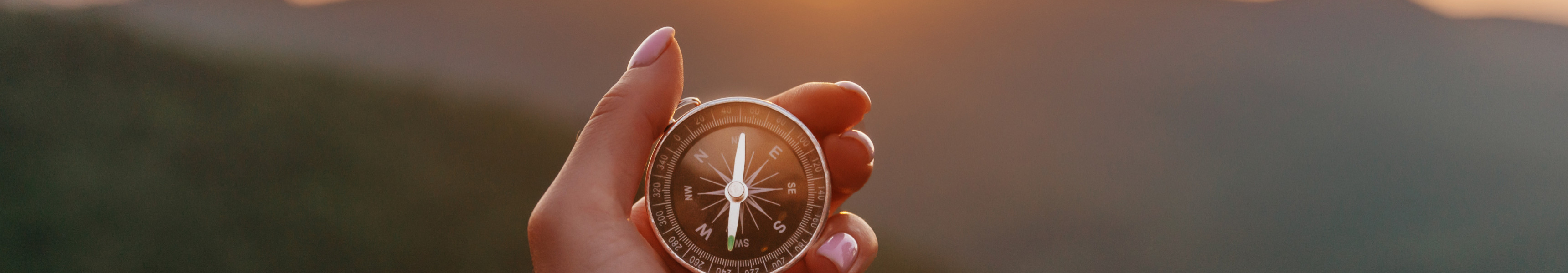 Hand hält einen Kompass vor Sonnenuntergang über einer Berglandschaft.
