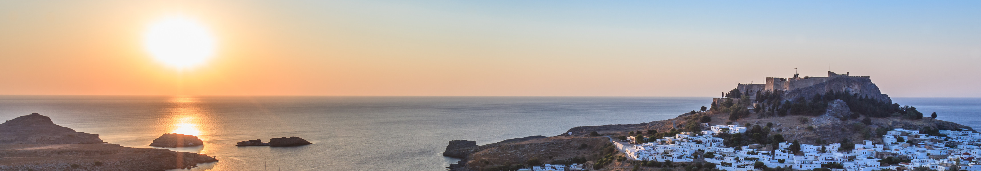Sonnenuntergang über der Küstenstadt Lindos auf Rhodos mit Bucht, weißen Häusern und Akropolis auf einem Hügel.