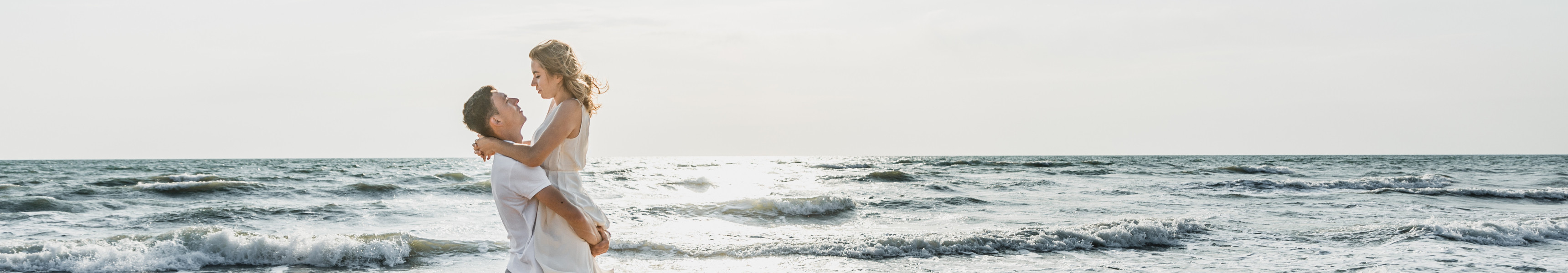 Ein Mann hebt eine Frau in weißem Kleid am Strand hoch, während beide lachend das Meer im Hintergrund und die untergehende Sonne genießen.