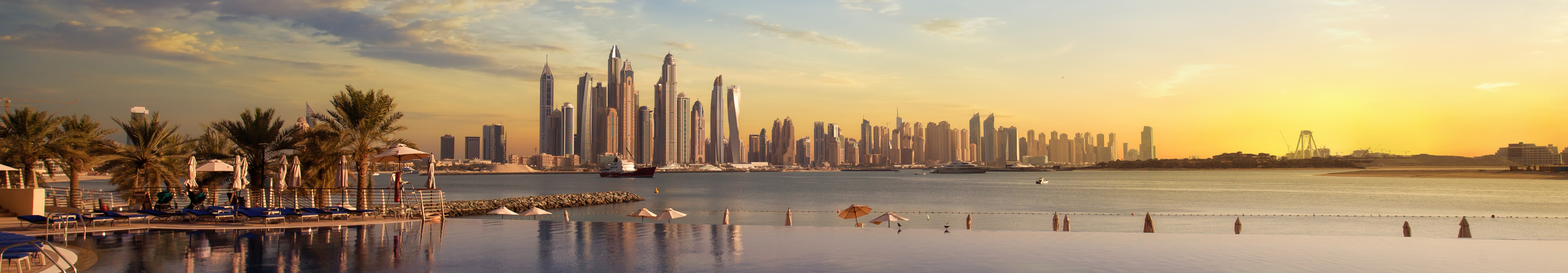 Infinity-Pool mit Palmen und Sonnenliegen im Vordergrund, dahinter das Meer und die Skyline von Dubai mit ihren modernen Hochhäusern bei Sonnenuntergang.