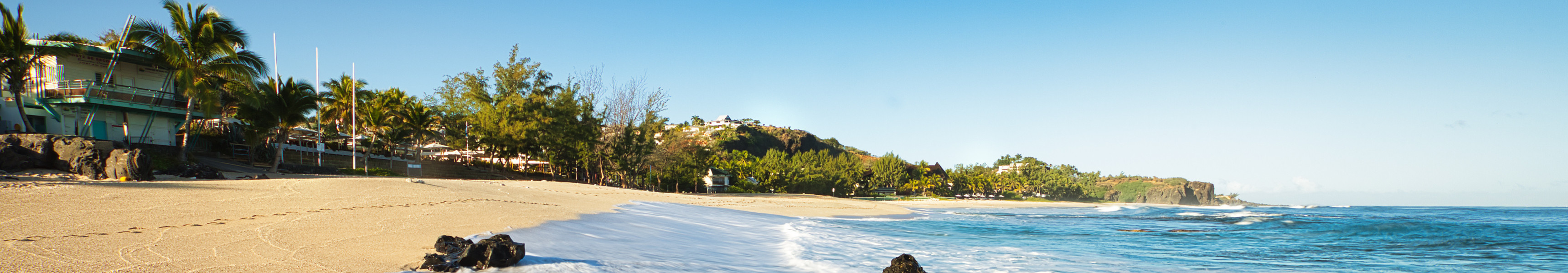 Wellen rollen über den goldenen Sandstrand von Saint-Gilles-les-Bains auf der Insel La Réunion, umgeben von Palmen und Küstenlandschaft unter klarem, blauem Himmel.