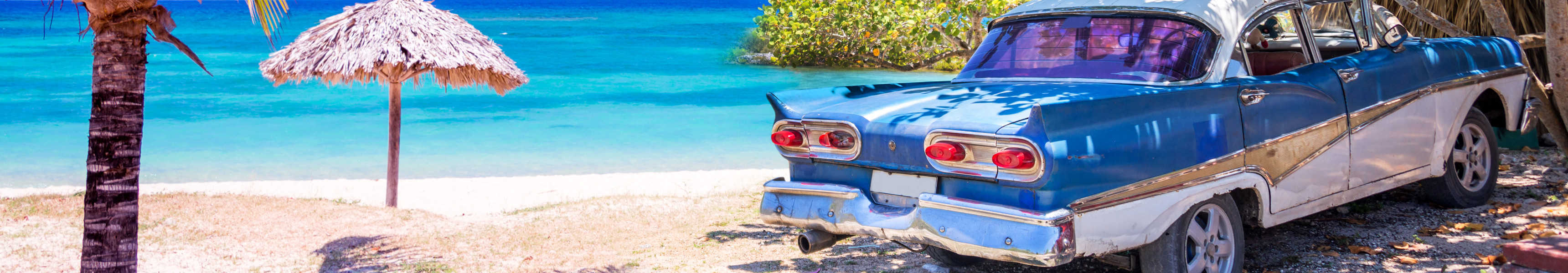 Ein klassisches Oldtimer-Auto steht im Schatten von Bäumen an einem tropischen Strand in Kuba, neben einer Palme und einer Strohhütte, mit türkisblauem Meer im Hintergrund.