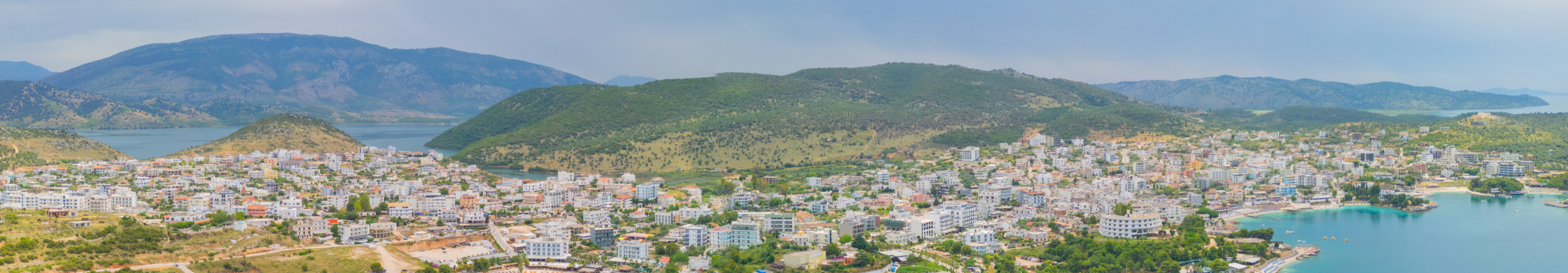 Panoramablick auf die Küstenstadt Saranda in Albanien mit zahlreichen Hotels, Stränden und türkisfarbenem Wasser, eingerahmt von grünen Hügeln und Bergen im Hintergrund.