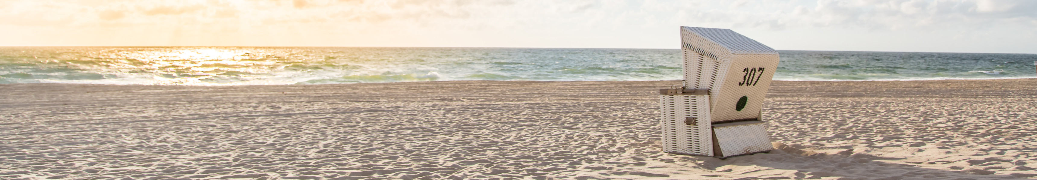 Ein einzelner Strandkorb mit der Nummer 307 steht am leeren Sandstrand von Föhr, während die Sonne hinter Wolken am Horizont über der Nordsee untergeht.