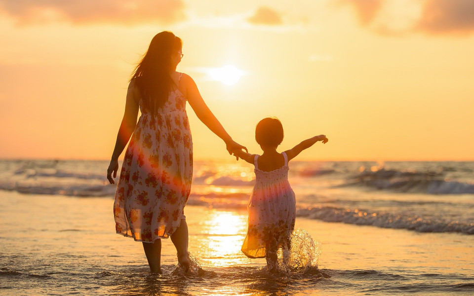Pauschalreisen buchen: Frau mit Kind spaziert am Strand bei Sonnenuntergang