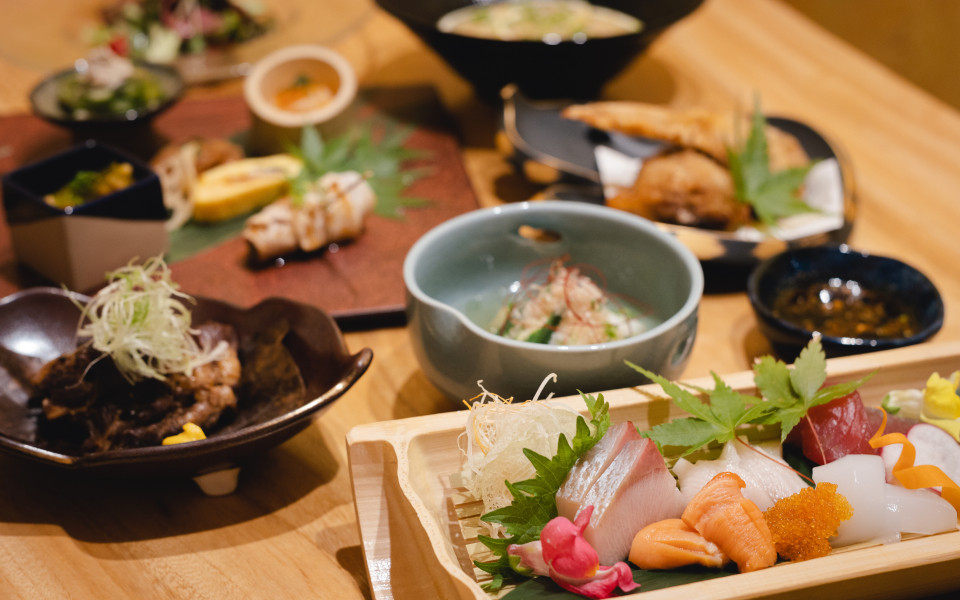 Japanisches Menü auf einem Holztisch mit kunstvoll angerichtetem Sashimi (Lachs, Thunfisch, weißer Fisch und Ikura) auf einem Holzbrett, dazu mehrere kleine Schalen mit Beilagen, Suppe und frittierten Speisen im Hintergrund.