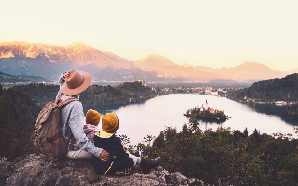 Mutter mit zwei kleinen Kindern sitzt auf einem Felsen und blickt auf den Bleder See in Slowenien &ndash; mit der Inselkirche im Wasser und schneebedeckten Bergen im Hintergrund bei Sonnenuntergang.